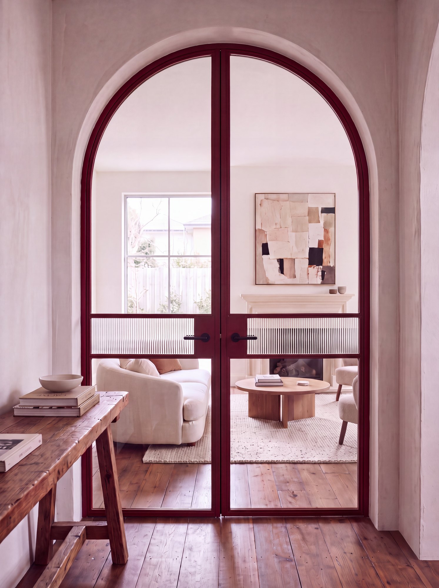 Oxblood red steel Crittall-style arched double doors with a reeded glass band, opening to a light living room in a New Zealand home