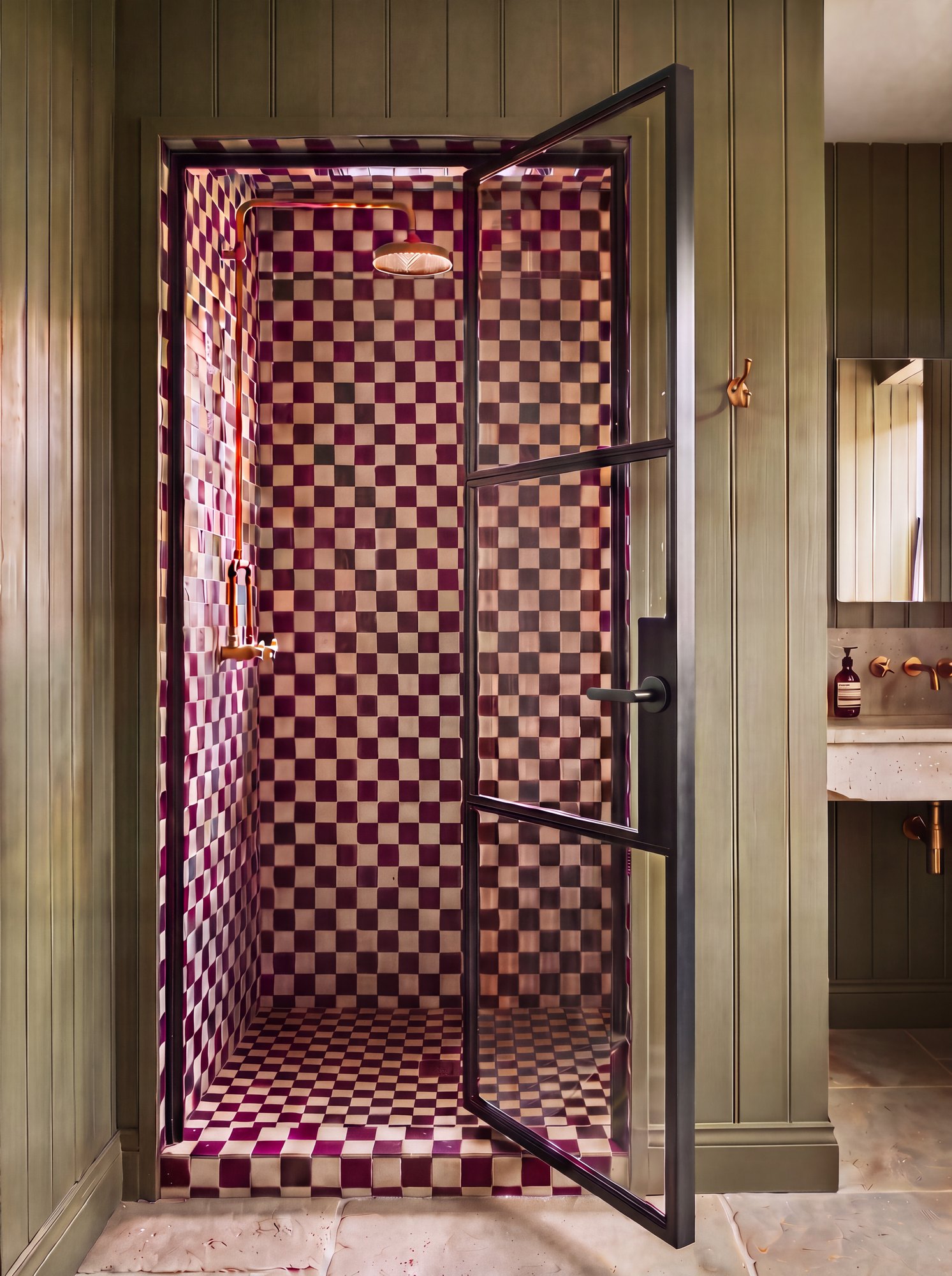 Black steel-framed glass shower door with a dummy lever handle, in an olive panelled bathroom with burgundy chequerboard tiles